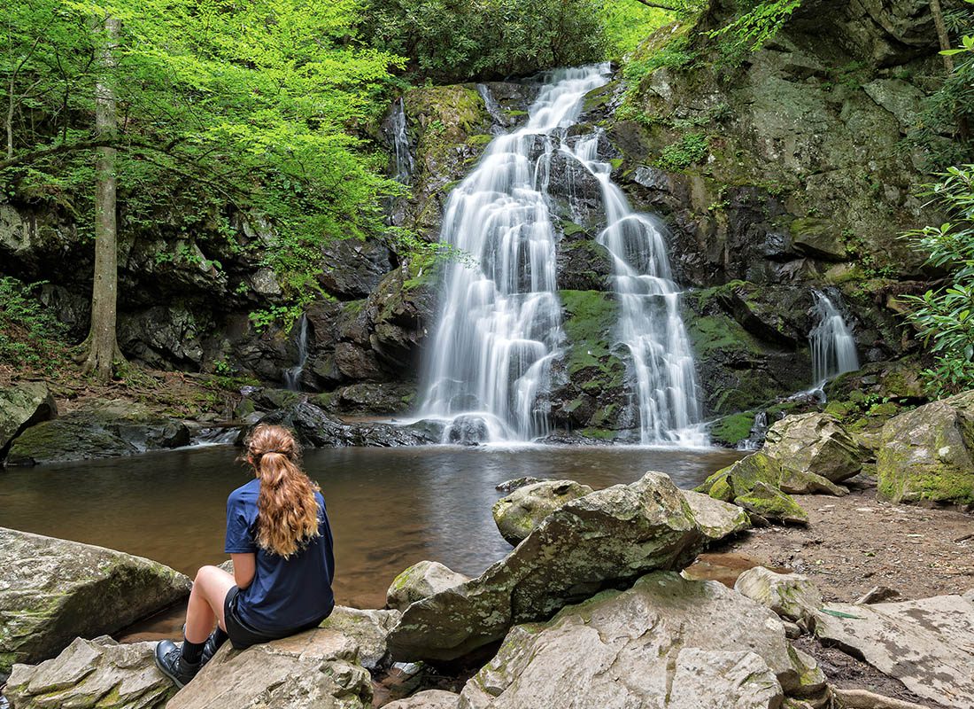 About Our Agency - Young Woman Sitting on Rocks as She Looks at a Waterfall in a Forest