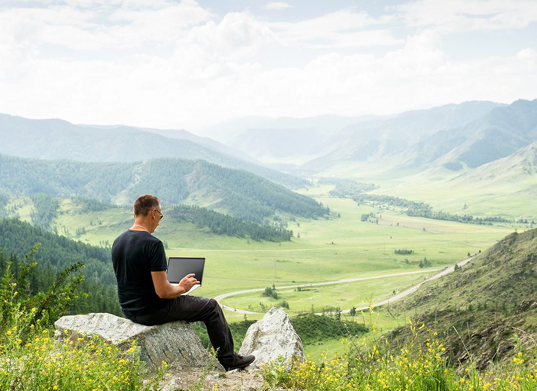 Blog - Man Sitting on a Large Stone in the Mountains Using a Laptop on a Sunny Day